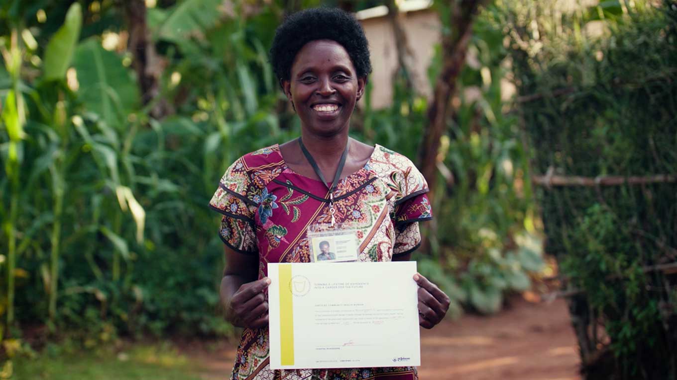 An African woman in a pink floral dress holds up a certificate, smiling.