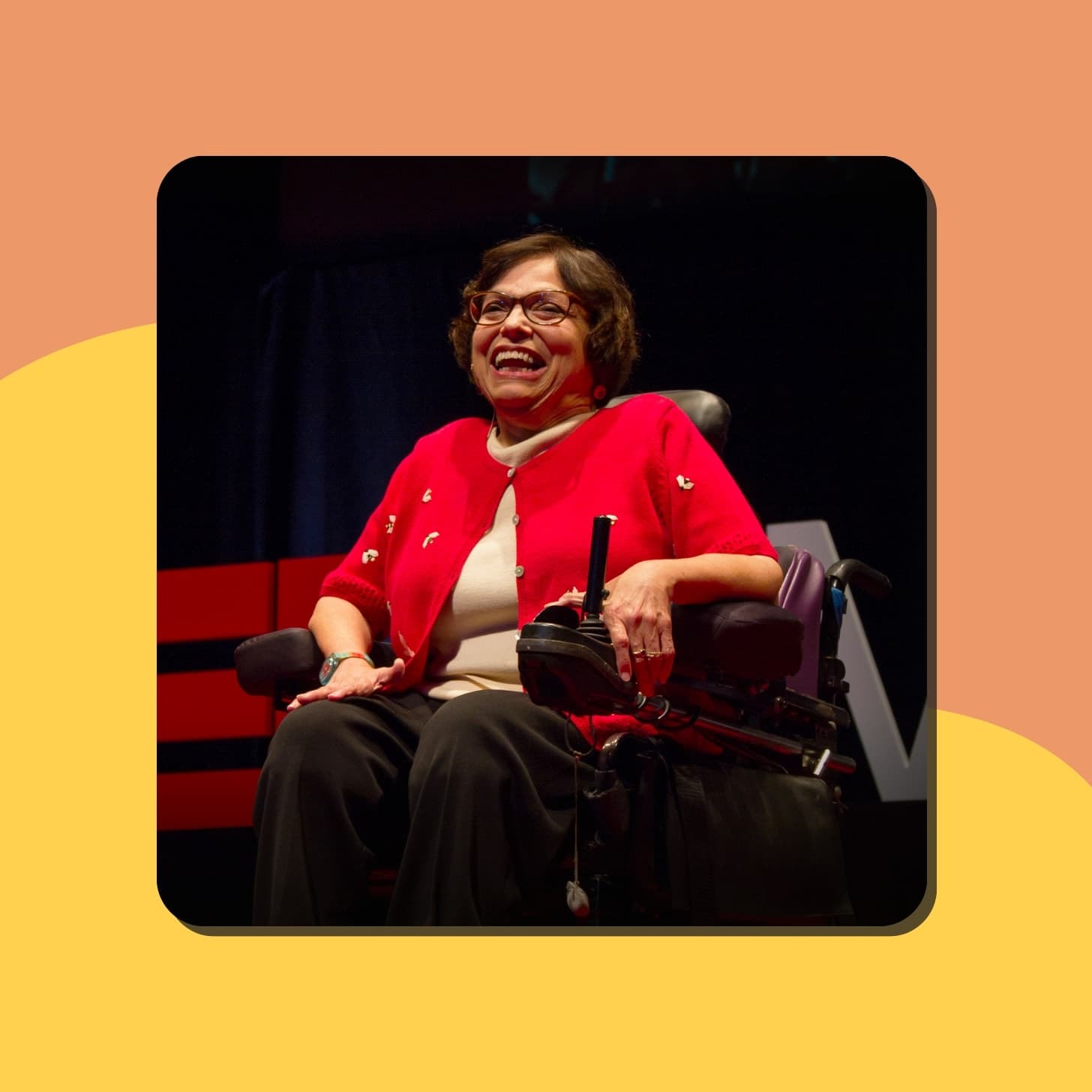 An older white woman with a beaming smile and cute glasses wears a red sweater and sits in a wheelchair on stage at a TED conference