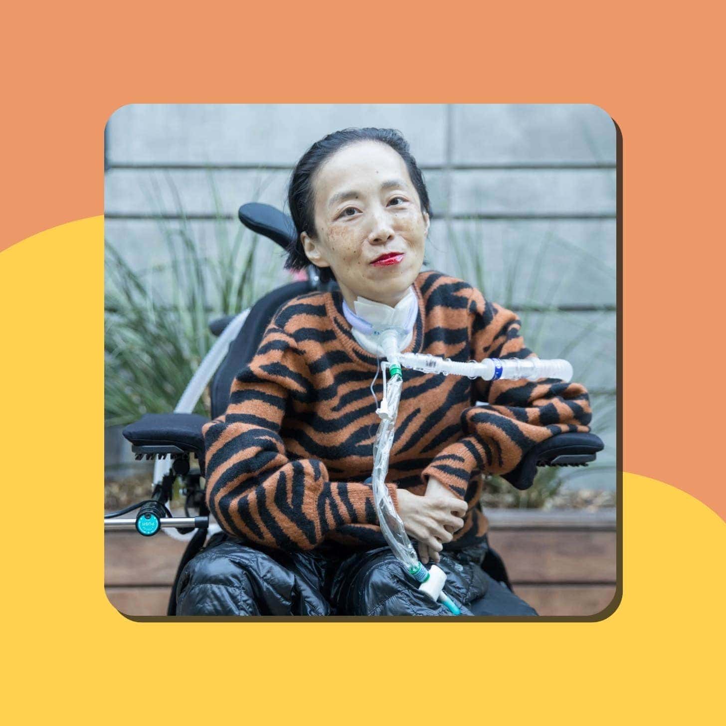 An Asian woman wears bold red lipstick and a fluffy orange sweater — she has a tube coming out of her neck and wrapping behind and in front of her as she sits in a wheelchair
