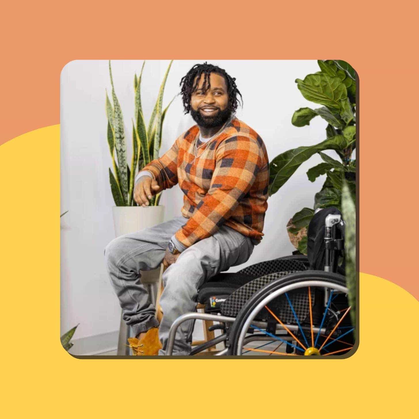 A Black man with cool hair, a beard, and a fashionable sweater, jeans, and boots smiles as he sits next to his wheelchair