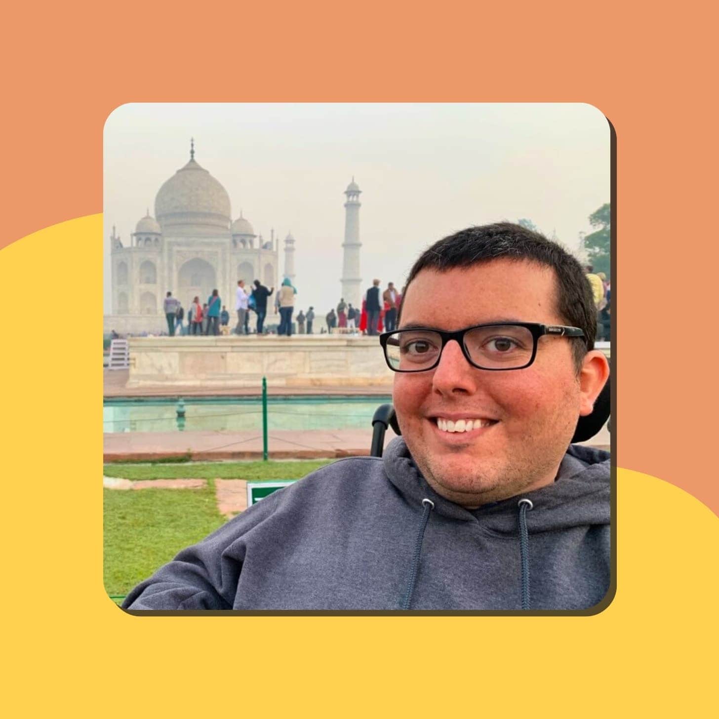 A white man smiling at the Taj Mahal, wearing glasses and sitting in a wheelchair