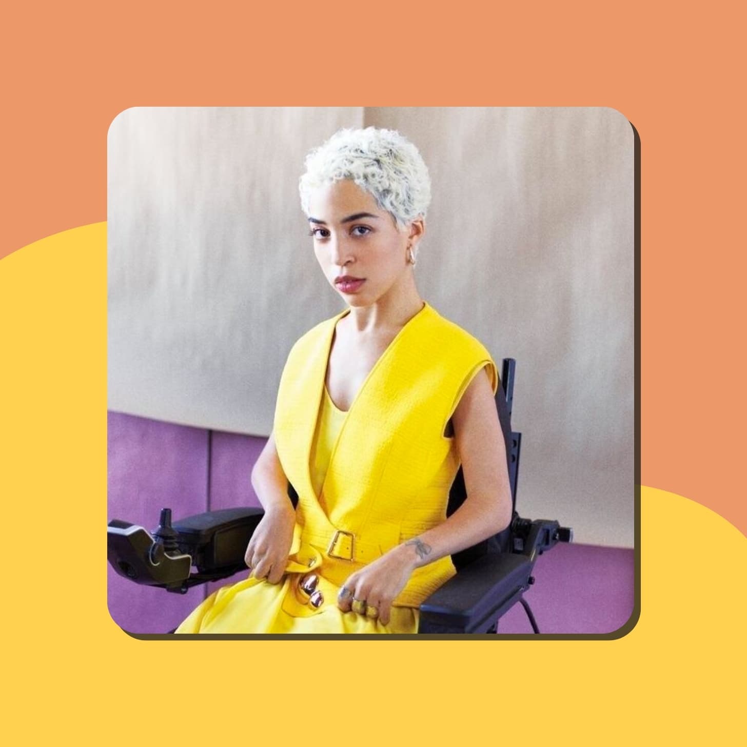 A woman with olive skin and bleached short hair wearing a fashionable yellow outfit, sitting in a wheelchair, in front of a photoshoot studio backdropr