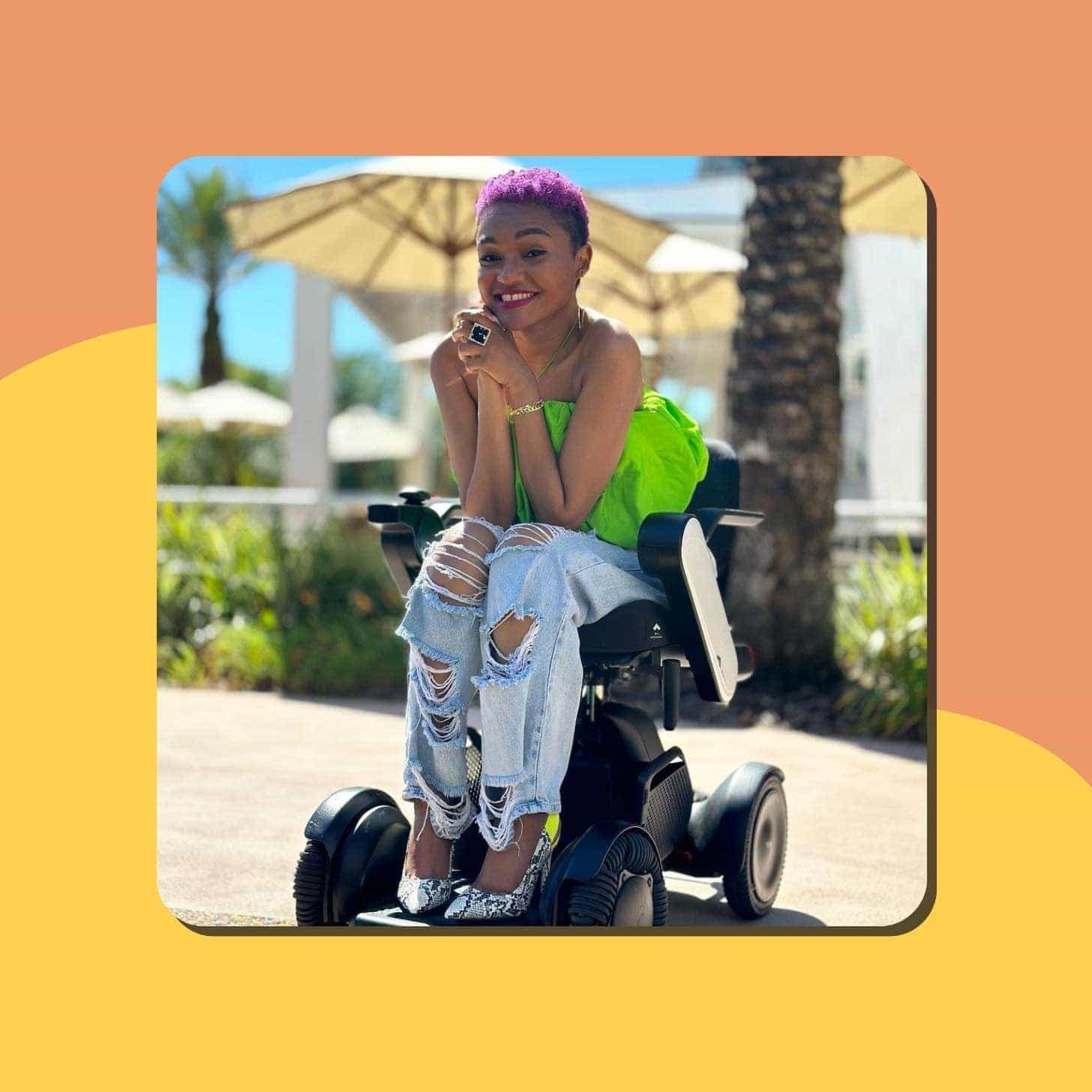 A Black woman with pink hair in trendy ripped jeans and a bright green top smiling in a wheelchair outdoors